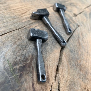 May include: Three silver metal hammer-shaped keychains on a wooden surface. The keychains are all different sizes and have a smooth, polished finish.