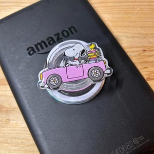 May include: A circular phone grip featuring a graphic of a white Snoopy character driving a pink convertible car. A small yellow Woodstock character sits atop a stack of colorful books in the back seat. The grip is attached to a black electronic device.