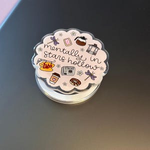 May include: A circular phone grip with a scalloped edge featuring a light pink background and the text "mentally in stars hollow". The design includes illustrations of a coffee pot, a gazebo, a newspaper, a donut, a coffee cup, and dragonflies.