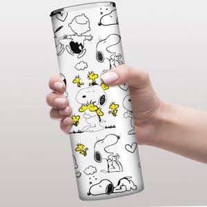May include: A white tumbler with a Peanuts theme.  The design features black and white illustrations of Snoopy and Woodstock in various poses.  Yellow accents highlight Woodstock. The tumbler is cylindrical and appears to be made of metal.