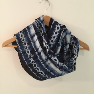 Peut inclure: Une écharpe en tie-dye bleu et blanc avec une bordure noire. L'écharpe est suspendue à un cintre en bois.