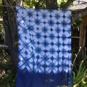 Peut inclure: Une écharpe en tie-dye bleu et blanc avec un motif géométrique. L'écharpe a une bordure frangée et est suspendue à une corde à linge.