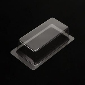 Puede incluir: Un embalaje de plástico transparente, rectangular, con tapa elevada. Diseñado para contener un producto, se muestra sobre un fondo negro. El embalaje es de plástico transparente.