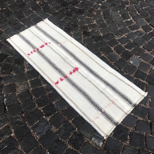 Puede incluir: Una alfombra rectangular blanquecina con rayas grises verticales y pequeños detalles rojos. La alfombra está colocada sobre una superficie oscura de adoquines, mostrando su diseño simple pero elegante. Ideal para un pasillo.