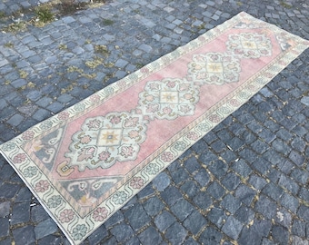 May include: A rectangular, vintage-style rug with a pink central field and intricate floral and geometric patterns. The rug has a border with floral motifs and is placed on a cobblestone surface. The rug's dimensions are not visible.