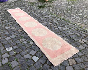 May include: A long, rectangular pink rug with a cream-colored border and repeating cream-colored floral designs. The rug is placed on a cobblestone surface, showcasing its length and pattern. The rug's dimensions are not visible.