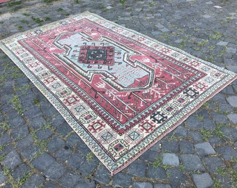 Puede incluir: Una alfombra rectangular vintage con un diseño geométrico central en tonos rojos, azul claro y negro. La alfombra presenta un borde detallado con patrones repetidos y está colocada sobre una superficie de adoquines.