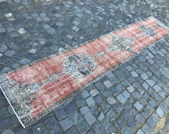 May include: A rectangular, distressed-look rug with a faded red and blue floral pattern. The rug has a long, narrow shape, ideal for a hallway or entryway. The rug's edges are finished with a white border, and it is placed on a cobblestone surface.