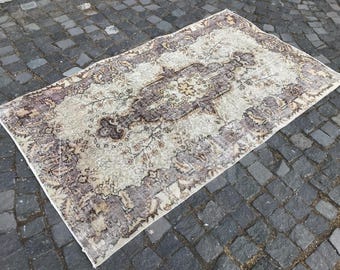 May include: A rectangular, vintage rug with a central medallion design. The rug has a cream-coloured field with a detailed, floral pattern in shades of brown and purple. It is placed on a cobblestone surface.