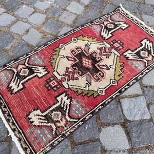 May include: A rectangular woven rug featuring a vibrant red background with a central cream-colored geometric medallion. The design includes stylized black and white bird-like motifs at the corners and edges, finished with short white fringe at ends.