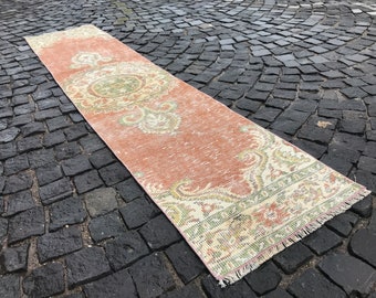 May include: A rectangular, distressed runner rug with a faded, vintage appearance. The rug features a salmon-colored field with ornate, cream-colored floral and scrollwork borders and a central medallion. The rug has a fringed edge.