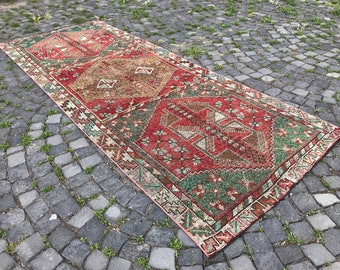 May include: A rectangular, vintage-style rug with a distressed appearance. The rug features a central design in shades of red, brown, and green, with geometric patterns and floral motifs. The rug is placed on a cobblestone surface.