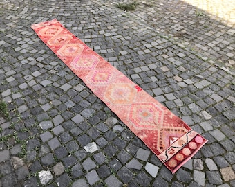 May include: A long, rectangular, vintage runner rug with a geometric pattern in shades of red, pink, and orange. The rug is placed on a cobblestone surface, showcasing its intricate design and warm color palette.