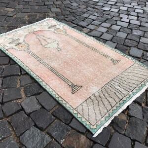 May include: Rectangular prayer rug with a light pink field and a decorative border. The rug features a central design with columns and an arch, and a fringe along the edges. The rug is placed on a cobblestone surface.
