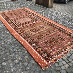 May include: A rectangular, hand-woven rug with an intricate geometric pattern. The rug features a central design in shades of brown, black, and red, framed by a border in a similar color palette. The rug is placed on a cobblestone surface.