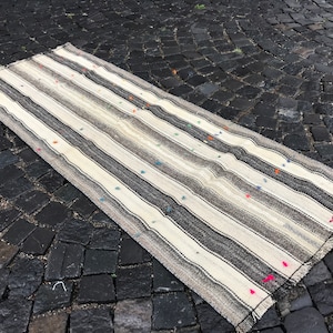 Puede incluir: Una alfombra rectangular a rayas con una paleta de colores neutros. La alfombra presenta bandas alternas de crema, gris y negro. Pequeños mechones de colores están dispersos por la superficie. La alfombra es probablemente de tela tejida.