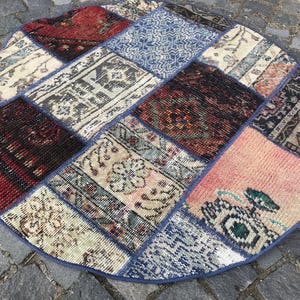 Könnte beinhalten: Ein runder Patchwork-Teppich mit blauem Rand. Der Teppich besteht aus verschiedenen rechteckigen und quadratischen Teilen in Rot-, Blau-, Beige- und Rosatönen. Das Design des Teppichs zeigt florale und geometrische Muster und schafft eine Vintage-Ästhetik.
