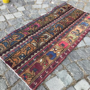 Könnte beinhalten: Handgewebter, rechteckiger Teppich mit Streifenmuster. Der Teppich zeigt eine Kombination aus Farben, darunter Burgunderrot, Braun, Blau und Rosa. Das Design integriert florale und geometrische Motive und schafft eine traditionelle Ästhetik. Der Teppich liegt auf einer Steinoberfläche.