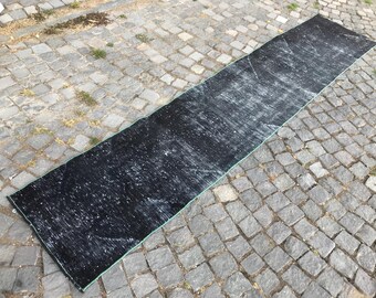 May include: A long, rectangular black rug with a distressed, faded appearance. The rug has a light green border and is placed on a cobblestone surface. The rug's design features white streaks.