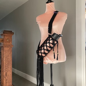May include: Black crocheted water bottle holder with a long fringe. The holder is worn on a mannequin with a pink shirt.