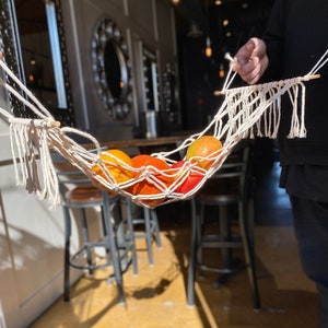 Puede incluir: Una hamaca de macramé blanca con cuatro naranjas y una manzana roja en su interior. La hamaca está colgada de una varilla de madera y está suspendida sobre un conjunto de sillas de metal.