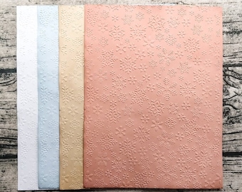 Puede incluir: Cuatro hojas de papel en relieve con un patrón de copo de nieve. Las hojas son blancas, azul claro, doradas y rosadas.