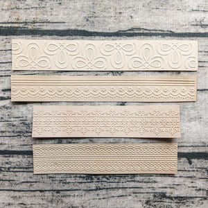May include: Four beige, rectangular embossing stamps with various decorative border designs. The stamps feature floral, scalloped, and geometric patterns. The stamps are arranged on a wooden surface with a natural grain.