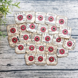 May include: A collection of small, square, off-white paper tags featuring vintage-style typography, numbers, and dates. Each tag has a circular red center with a brass-colored metal eyelet reinforcement. The tags are arranged in a cluster.