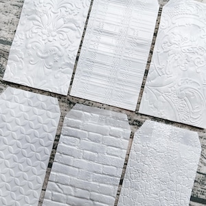 Puede incluir: Seis etiquetas de regalo blancas con patrones en relieve. Los patrones incluyen diseños florales, geométricos y de ladrillo. Las etiquetas están dispuestas sobre una superficie de madera, mostrando sus texturas y detalles.