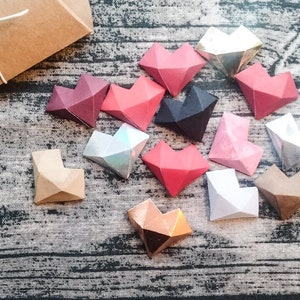 May include: A collection of 12 origami hearts in various colours, including red, pink, brown, black, silver, and gold. The hearts are arranged on a textured surface.