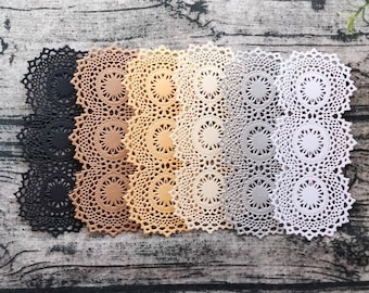 May include: Seven lace doilies in various colors, including black, brown, beige, white, and gray. The doilies are arranged in a row on a wooden surface.
