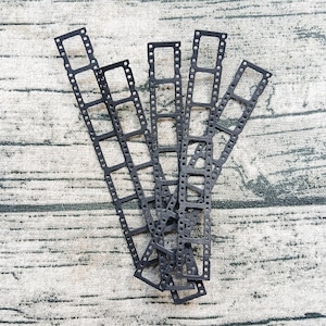 May include: Four black, die-cut strips resembling vintage 35mm film negatives. Each strip features a series of rectangular frames with small, circular sprocket holes along both vertical edges, arranged in a fan-like pattern on a textured surface.