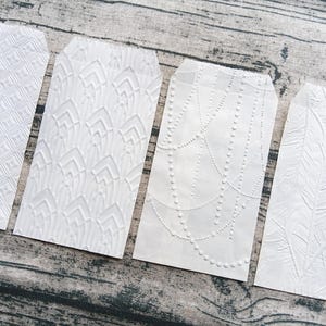 Op de afbeelding: Vier witte cadeaulabels met reliëfpatronen. De labels hebben verschillende ontwerpen: een geometrisch ruitpatroon, een gestileerde waaier, een kralenkettingontwerp en een varenachtig patroon. De labels zijn gerangschikt op een houten oppervlak.