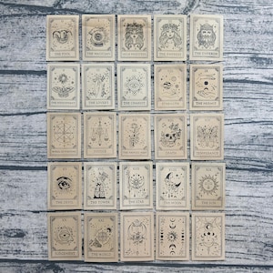 May include: A collection of twenty-five rectangular tarot cards arranged in a grid, each featuring black line art illustrations on a light tan background. The cards display various mystical symbols, figures, and celestial motifs with labels.