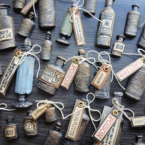 Puede incluir: Una colección de frascos de boticario en miniatura con acabados oscuros y desgastados, cada uno con etiquetas de estilo vintage con textos como Bone Powder, Poison y Witch Hazard. Muchos frascos tienen pequeñas etiquetas de papel atadas con hilo blanco.