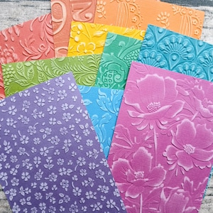 Op de afbeelding: Een verzameling kleurrijke, getextureerde papieren vellen met reliëf bloemen- en patroonontwerpen. De vellen zijn in verschillende kleuren, waaronder roze, paars, blauw, groen, oranje en geel. De ontwerpen omvatten bloemen, bladeren en andere decoratieve elementen.