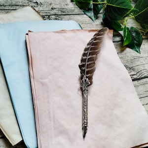 May include: A stack of three sheets of paper, one white, one light blue, and one light pink, with a silver and brown feather quill pen on top of the pink sheet.