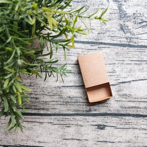 May include: A small, rectangular, empty cardboard matchbox with a light brown, natural finish. The box consists of an outer sleeve and an inner sliding tray, both made of plain, unprinted, tan-colored paperboard material.