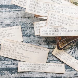 May include: A selection of aged paper strips featuring Bible verses printed in black ink. The strips are arranged on a blue and white patterned fabric. A small glass container is visible.