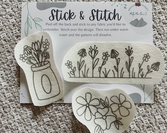 May include: Three black and white embroidery patterns on white fabric. The patterns are of flowers in a jar, a row of flowers, and a cluster of flowers. The text "Stick & Stitch" is visible on the card behind the patterns. Peel off the back and stick to any fabric you'd like to embroider. Stitch over the design. Then run under warm water and the pattern will dissolve.
