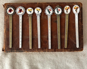 Puede incluir: Ocho marcapáginas de metal con diseños florales bordados. Cada marcapáginas tiene una regla en la parte posterior. Los marcapáginas están dispuestos en una fila sobre un libro encuadernado en cuero marrón.