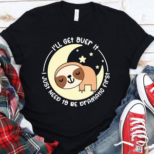 Puede incluir: Camiseta negra con un gráfico blanco de un perezoso durmiendo en una luna creciente. El texto "I'll get over it I just need to be dramatic first" está escrito alrededor de la luna.