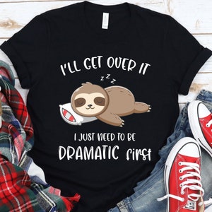 Puede incluir: Camiseta negra con un perezoso de dibujos animados durmiendo sobre una almohada. El texto dice "I'll get over it, I just need to be dramatic first" en blanco. El perezoso es marrón con la cara color crema y está sobre una almohada blanca con rayas rojas.
