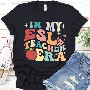 Könnte beinhalten: Schwarzes T-Shirt mit einem bunten Retro-Design, auf dem "In My ESL Teacher Era" steht. Das Design zeigt einen roten Apfel, einen Bleistift und Sterne.