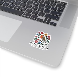 May include: A white sticker featuring a bird perched on a branch, surrounded by colorful flowers. The sticker has the text "RADICALIZED BY BASIC DECENCY" in a black, stylized font. The sticker is placed on a laptop.