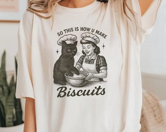 Può includere: T-shirt color crema con un'illustrazione in stile vintage. La grafica raffigura una donna e un gatto nero con cappelli da chef, che mescolano ingredienti in una ciotola. Il testo recita "So this is how u make Biscuits."
