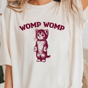 Puede incluir: Una camiseta blanca con un gráfico de un gato marrón y el texto "WOMP WOMP" impreso en la parte delantera.