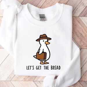 Może przedstawiać: Biała bluza z kapturem z kreskówkową kaczką w brązowym kapeluszu i teczką. Pod spodem wydrukowano napis "LET'S GET THE BREAD". Bluza wykonana jest z miękkiego materiału.