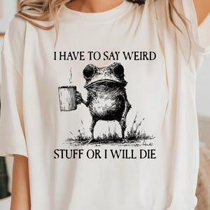 Puede incluir: Camiseta color crema con una ilustración en blanco y negro de una rana sosteniendo una taza. El texto encima de la rana dice "I HAVE TO SAY WEIRD", y debajo, "STUFF OR I WILL DIE".