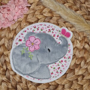 Könnte beinhalten: Ein grauer Filz-Elefanten-Applik mit einer rosa Blume und einem Schmetterling. Der Elefant ist von einem weißen Hintergrund mit rosa Blumen umgeben.